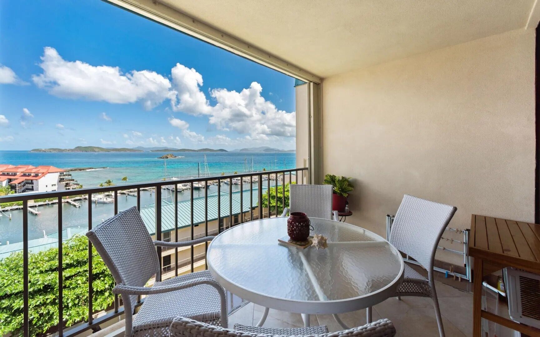Un balcone con un tavolo rotondo, due sedie in vimini, una pianta in vaso e vista sul mare e sugli edifici.