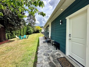 A house with a stone pathway leading to a front door, a small front yard with a lawn and chairs, and a large tree to the left. - Beautiful new 1-bed studio suite with AC & big private yard in charming Burnaby (Burnaby)