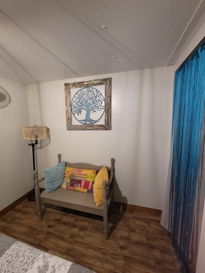 Ein Zimmer mit Holzboden, einer Couch mit Kissen, einem gerahmten Bild von einem Baum, einer Stehlampe und einem blauen Vorhang.