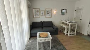 A living area with a grey sofa, a small white table with a woven basket, a dining table with stools, and a wall-mounted mirror. - Casa DiLauNa - Cozy apartment in the heart of Albufeira - Portugal (Albufeira)