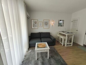 Ein Wohnbereich mit grauem Sofa, einem kleinen weißen Tisch mit einem gewebten Korb, einem Esstisch mit Hockern und einem wandmontierten Spiegel.