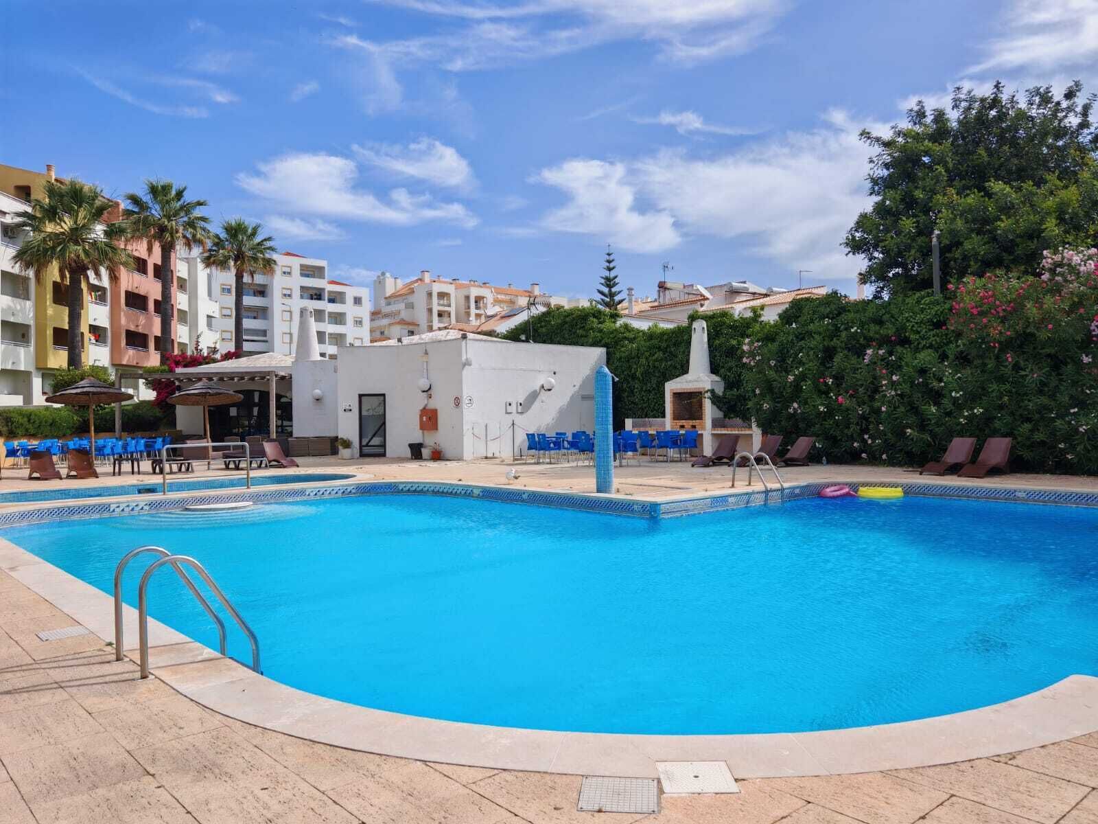A swimming pool with lounge chairs and umbrellas, surrounded by buildings and greenery.