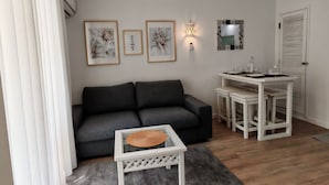 A living room with a dark grey sofa, a white coffee table with a woven placemat, a small white table with stools, and wall-mounted artwork. - Casa DiLauNa - Cozy apartment in the heart of Albufeira - Portugal (Albufeira)