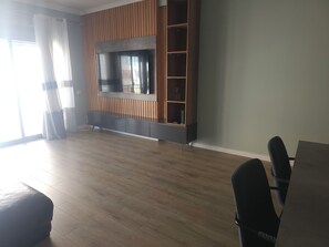 Ein geräumiges Zimmer mit großem Flachbildfernseher an einer Wand aus Holzpaneelen, einem Esstisch mit Stühlen und einem großen Fenster mit leichtem Vorhang.