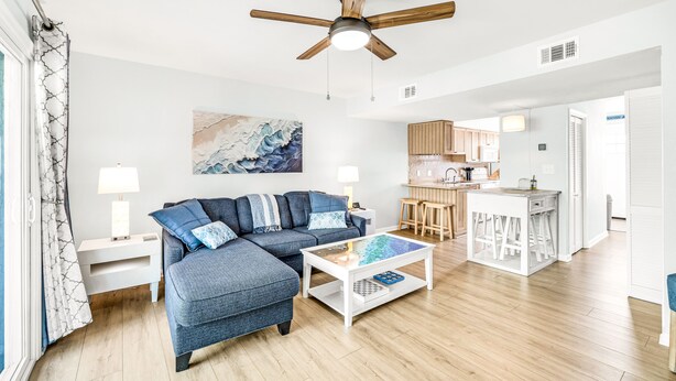 Ein modernes Wohnzimmer mit Ecksofa, Couchtisch, Deckenventilator und einer Küchenzeile im Hintergrund.