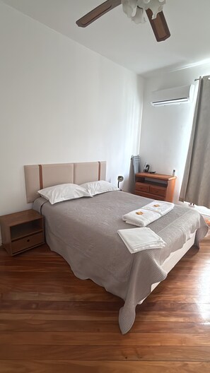 A neatly made bed with a headboard, a bedside table, a wooden nightstand, a wall-mounted air conditioner, and a window with curtains. - Apartment with garage, across from the subway and with a view of Christ the Redeemer. (Rio de Janeiro)