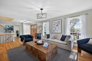 Ein geräumiges Wohnzimmer mit einem Holztisch, zwei Sofas, einer Treppe und einem großen Fenster mit Vorhängen.
