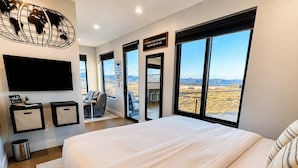 A modern hotel room with a large bed, a flat-screen TV, a world map artwork, and a view of the desert landscape. - The Dark Sky Haven, Luxe Casita, Views, Sleeps 2 (Page)