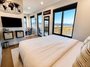 A modern hotel room with a large bed, a flat-screen TV, a world map artwork, and a view of the desert landscape. - The Dark Sky Haven, Luxe Casita, Views, Sleeps 2 (Page)