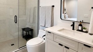 A modern bathroom with a glass shower enclosure, a white toilet, a sink with a mirror, and a small stool. - The Dark Sky Haven, Luxe Casita, Views, Sleeps 2 (Page)