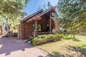 A rustic log cabin with a porch, surrounded by trees and a gravel path.