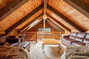 A cozy attic room with a wooden ceiling, a large window, a wooden table, leather sofas, and a fluffy rug.