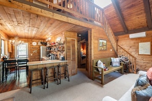 A wooden cabin interior with a kitchen, dining area, and a staircase leading to a loft.