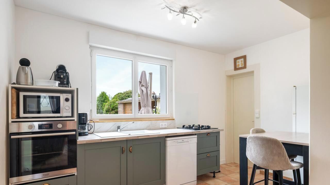 Eine moderne Küche mit Mikrowelle, Backofen und Kaffeemaschine auf der Arbeitsfläche, einem Fenster mit Blick ins Grüne und einem Essbereich mit einem Stuhl.