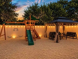 A playground with a slide and swings, a covered seating area, and a fence with string lights. - Wilton Manors • Las Olas • Beach • Downtown FTL (Fort Lauderdale)