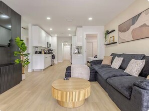 A modern living room with a dark grey sofa, a round wooden coffee table, and a kitchen in the background. - Wilton Manors • Las Olas • Beach • Downtown FTL (Fort Lauderdale)