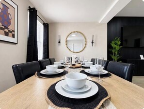 A dining area with a wooden table set for six, black chairs, a mirror, and a black wall with a television. - Wilton Manors • Las Olas • Beach • Downtown FTL (Fort Lauderdale)