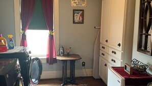 A laundry room with a washing machine, a drying rack, a small table with a lamp, a red and blue patterned rug, and a sign indicating laundry services.