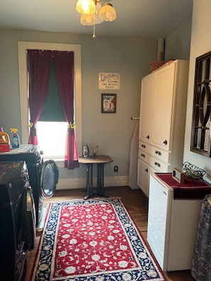 Une buanderie équipée d’un lave-linge, d’un étendoir, d’une petite table avec une lampe, d’un tapis aux motifs rouges et bleus, et d’un panneau indiquant les services de blanchisserie.