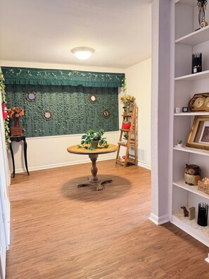 A room with a wooden floor, a round table with a vase of flowers, a green wall with decorative mirrors, and a bookshelf with various items.