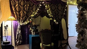 A room with a wooden floor, a table with a vase of flowers, a chair, a mirror, and draped curtains. - Victorian Fairy Cottage (Houston)