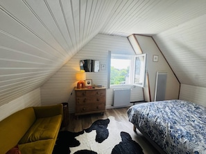 Ein Schlafzimmer mit schrägen Wänden, einer Holzkommode, einem Bett mit gemustertem Überwurf, einem gelben Sofa und einem Kuhfellteppich.