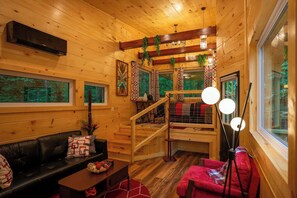 Innenansicht einer Holzhütte mit einer Couch, einem kleinen Tisch, einer Treppe und einem Fenster mit Blick auf Grünflächen.