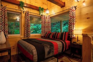 A cozy wooden cabin bedroom with a bed, curtains, a nightstand, and a window with a view of greenery. - Terrace Creek Retreat | Creekside Hot Tub Escape | 5 Min From Nada Tunnel! (Stanton)
