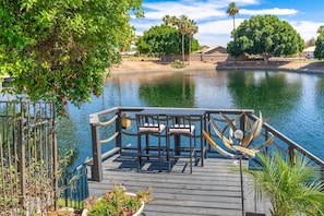 A wooden dock with chairs and a table, overlooking a calm body of water with trees and buildings in the background. - Lakefront Oasis | Infinity Edge Pool & Golf Views (Glendale)