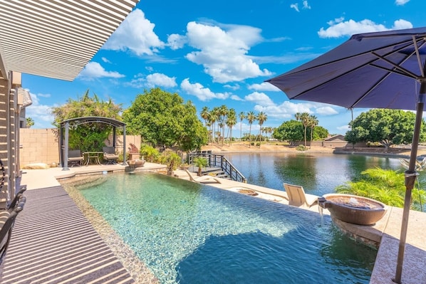 A swimming pool with a pergola, lounge chairs, and a view of a lake with palm trees. - Lakefront Oasis | Infinity Edge Pool & Golf Views (Glendale)