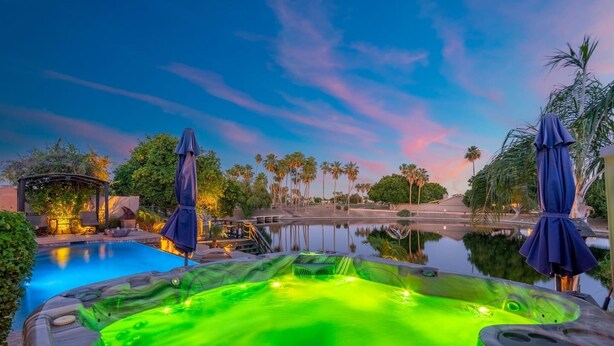 A hot tub with a green light, surrounded by palm trees and a pool, under a twilight sky. - Lakefront Oasis | Infinity Edge Pool & Golf Views (Glendale)