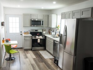 Eine moderne Küche mit Edelstahlgeräten, einer Mikrowelle, einem Backofen und einem Kühlschrank. Ein Fenster mit Jalousien, ein Waschbecken und ein kleiner Hocker sind ebenfalls vorhanden.