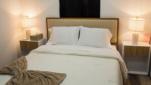 Ein ordentlich bezogenes Bett mit weißen Leinentüchern, zwei Nachttischlampen, ein Nachttisch mit Taschentuchbox und eine braune Tagesdecke.