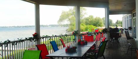A covered outdoor dining area with a view of a lake, featuring a long table with chairs and potted plants.