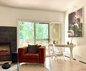 Ein Zimmer mit Kamin, einem roten Sessel, einem weißen Schreibtisch, einem transparenten Stuhl und einem großen Fenster mit Blick auf Bäume.