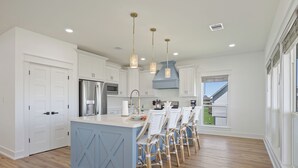 Una cocina moderna con armarios blancos, una isla central, luces colgantes y vistas al exterior.