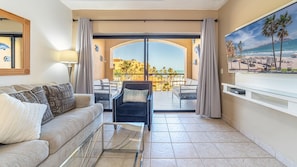 Ein Wohnzimmer mit Sofa, Sessel und Glastisch. Ein Balkon bietet Blick auf den Strand und Palmen.