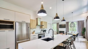 A modern kitchen with stainless steel appliances, a central island with a sink, pendant lighting, and a dining area with a view. - Astral - Private Hot Tub, Pet-Friendly (Pont-Rouge)