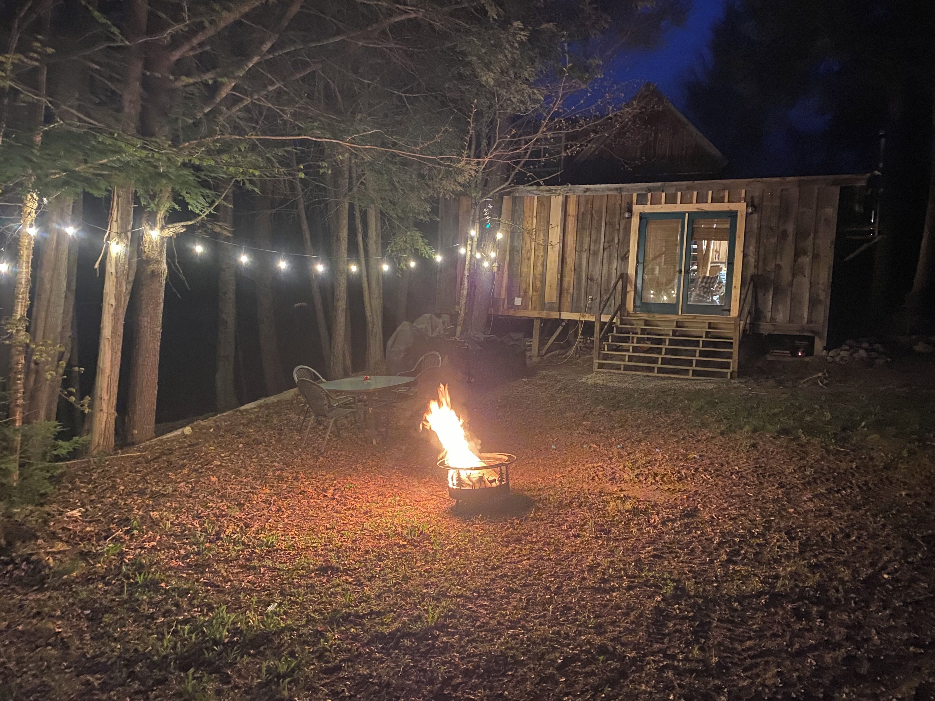 Une cabane en bois rustique, avec un foyer au milieu de la cour, entourée d’arbres et illuminée de guirlandes lumineuses.
