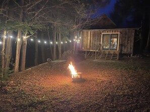 A rustic wooden cabin with a fire pit in the yard, surrounded by trees and string lights.