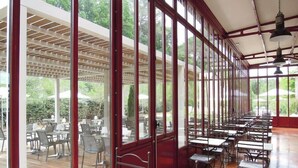 A covered outdoor dining area with tables and chairs, large windows, and a wooden floor. - Hotel De L'estelou (Sommieres)