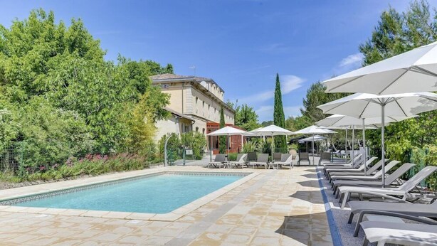 A swimming pool with lounge chairs and umbrellas, surrounded by greenery and a building. - Hotel De L'estelou (Sommieres)