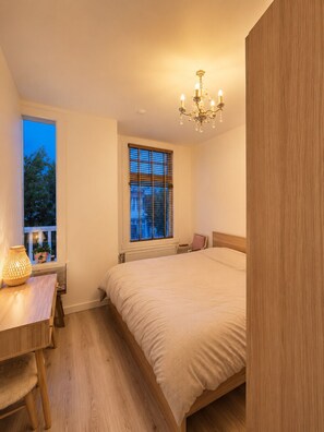 Ein Schlafzimmer mit einem Bett, einem Fenster mit Jalousien, einem Nachttisch mit Lampe und einem Holzboden.