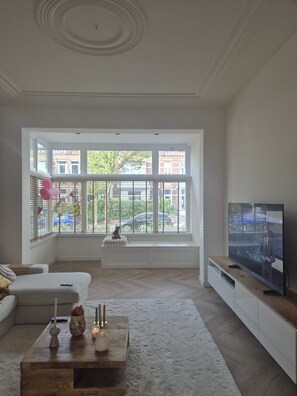 Ein geräumiges Wohnzimmer mit großem Fenster, Fernseher und einem Holztisch.