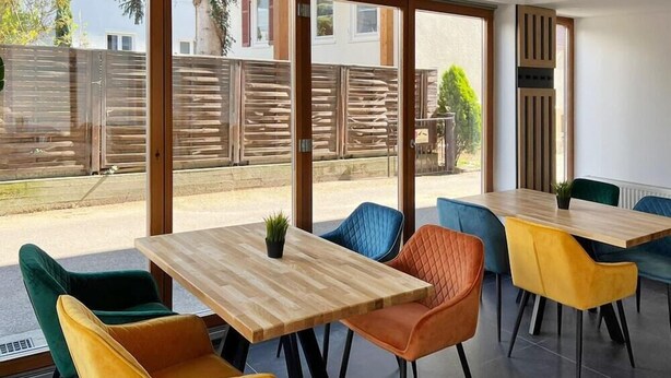Un espace de restauration moderne avec des tables en bois et des chaises colorées, de grandes fenêtres donnant sur une clôture et de la verdure.