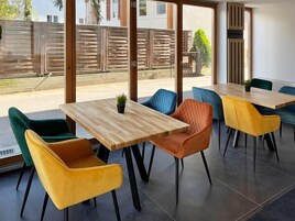 A modern dining area with wooden tables and colorful chairs, large windows with a view of a fence and greenery.