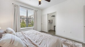 A bedroom with a bed, a ceiling fan, a window with curtains, a walk-in closet, and a view of buildings outside. - Frisco Getaway with Pool, Gym & Free Parking (Frisco)