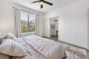 A bedroom with a bed, a ceiling fan, a window with curtains, a walk-in closet, and a view of buildings outside. - Frisco Getaway with Pool, Gym & Free Parking (Frisco)