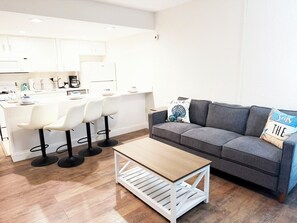 Une cuisine moderne avec des meubles blancs, un coin repas avec des chaises blanches, un canapé gris agrémenté de coussins décoratifs et une table basse en bois.