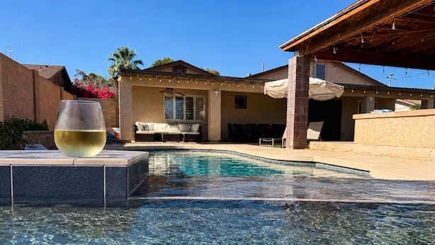 Una piscina cristalina con una copa de vino blanco sobre un repisa, un patio cubierto y un edificio con fachada color beige al fondo.